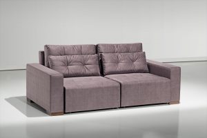 A imagem mostra um espaço com uma parede branca e um piso mais escuro que reflete o sofá. No centro da imagem encontra-se o sofá Retrátil Jhow na cor marrom e com detalhes em seu encosto.