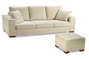 A imagem mostra um ambiente com fundo totalmente branco. No centro encontra-se o Sofá Spazio, na cor bege com pés de madeira nos cantos do sofá. Na frente está um puff quadrado com pés de madeira e nas mesmas cores do sofá.