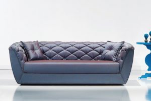 A imagem mostra um ambiente claro, com parede branca e piso branco. No centro encontra-se um sofá na cor azul com detalhes no estofado e dois travesseiros um em cada canto do sofá também nas cores azuis. Na lateral direita do sofá temos a metade de uma mesinha azul.