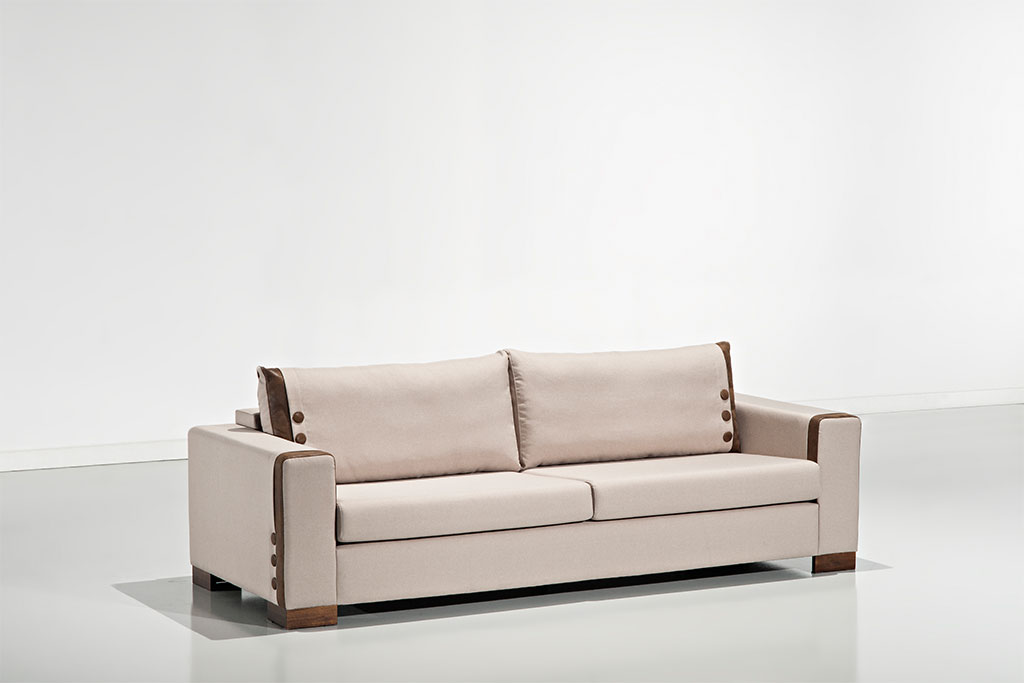 A imagem mostra um espaço claro com paredes e piso branco, refletindo o sofá. No centro da imagem está o Sofá Accord, na cor branca com detalhes em marrom e pés em madeira.