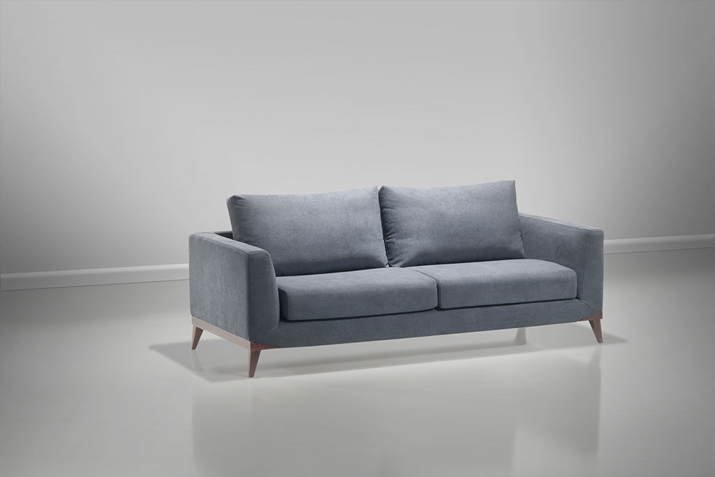 : A imagem mostra um espaço com parede e piso brancos. No centro da imagem encontra-se o sofá Kari, na cor azul claro, seus pés são de madeira.