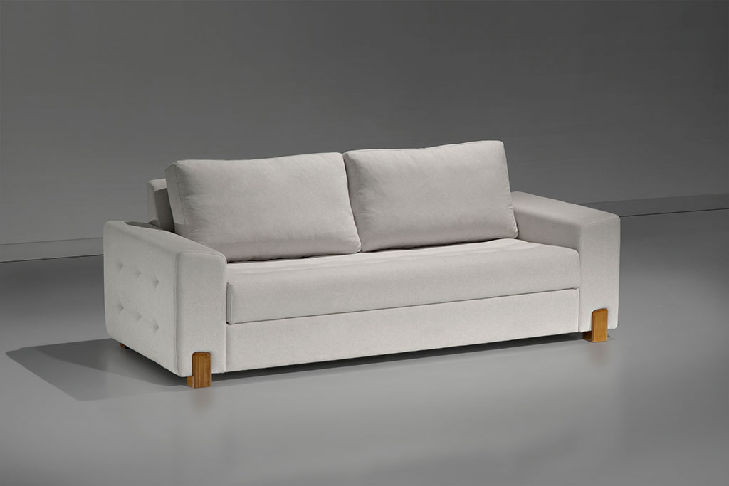 A imagem mostra um espaço escuro. Ao centro encontra-se o sofá Lazio, na cor branca, com alguns detalhes no estofado e pés em madeira.