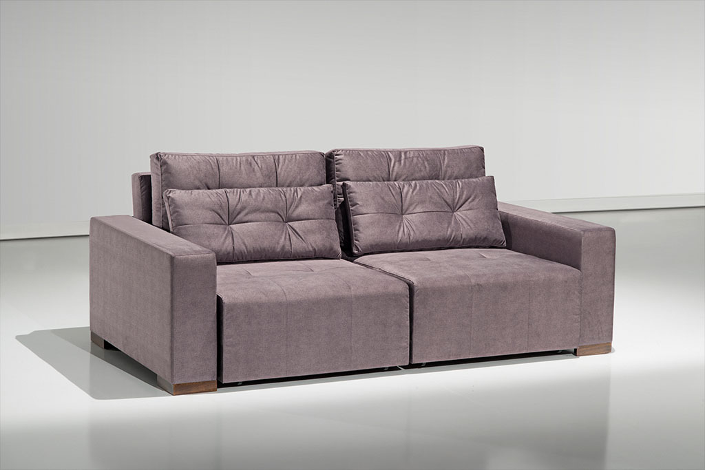 A imagem mostra um espaço com uma parede branca e um piso mais escuro que reflete o sofá. No centro da imagem encontra-se o sofá Retrátil Jhow na cor marrom e com detalhes em seu encosto.