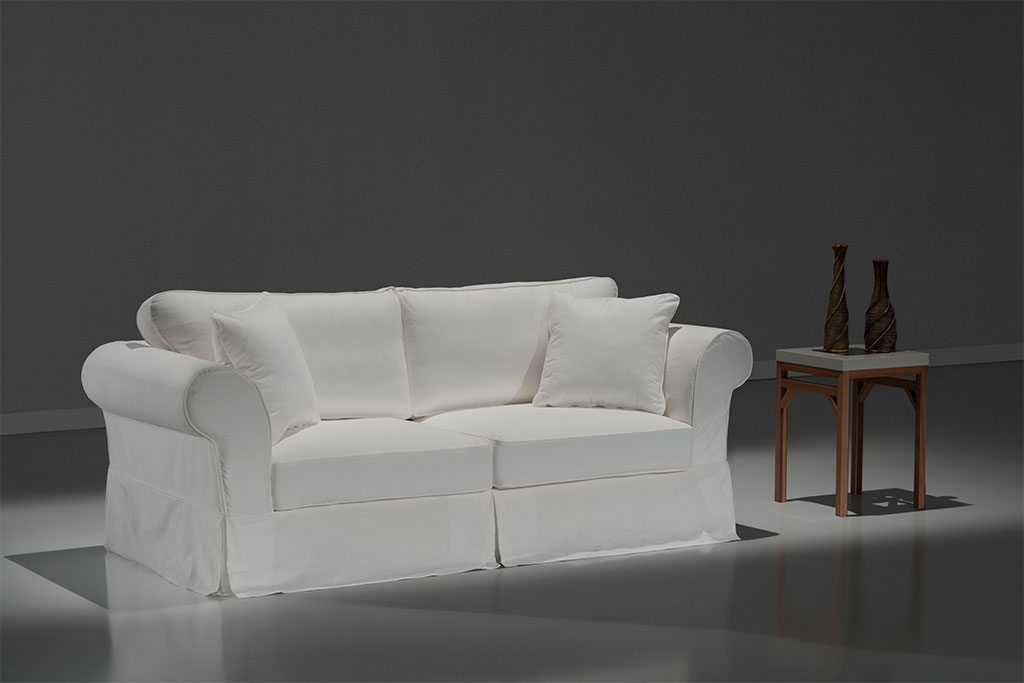A imagem mostra um espaço com fundo cinza e piso branco. No centro encontra-se um sofá usando a Capa Liege na cor branca. Ao lado direito do móvel há uma mesa marrom e sobre ela estão dois decorativos na cor marrom.