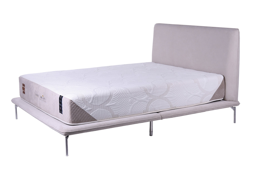A imagem mostra uma cama, em ângulo diagonal, com a cabeceira e parte inferior estofadas na cor cinza claro. Na base da cama há um colchão branco com uma textura em relevo. Os pés da cama são finos e de alumínio. O fundo da imagem é branco.