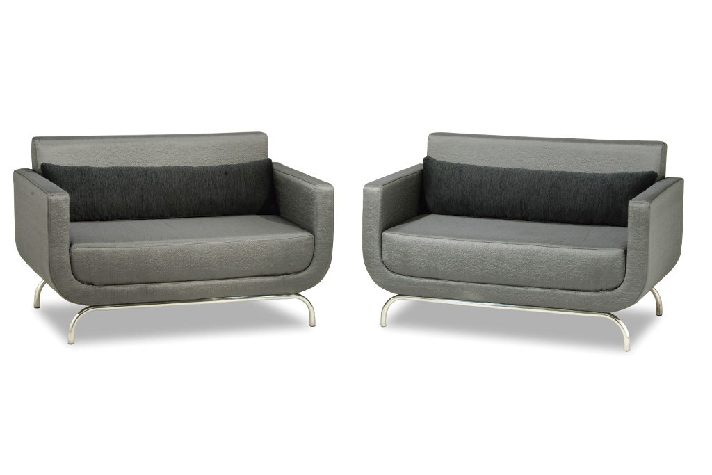 A imagem mostra duas poltronas iguais, em cor cinza, uma ao lado da outra. Elas possuem os pés em aço inox. As duas tem uma almofada decorativa preta sobre o assento. O fundo da imagem é branco.