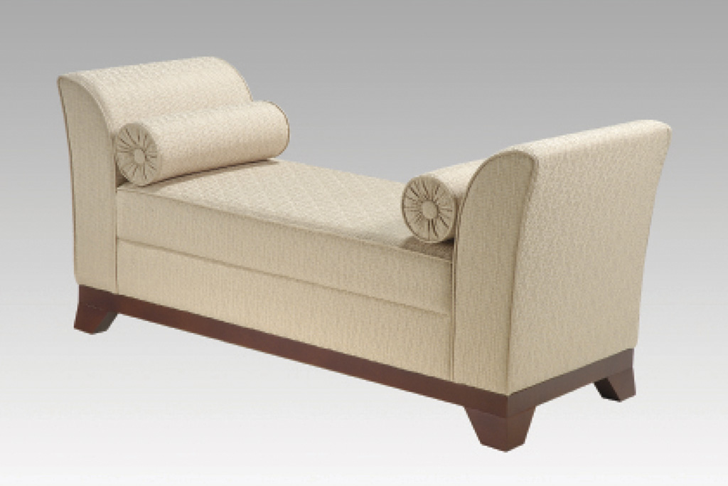 A imagem mostra o sofá Karol na cor bege com uma textura. Ele possui dois rolinhos decorativos próximo aos braços do sofá, um de cada lado. A parte inferior do sofá é de madeira, em um tom marrom escuro. O fundo da imagem é cinza claro.