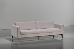 A imagem mostra um ambiente claro, com paredes brancas e piso refletindo partes do sofá. No centro da imagem encontra-se um sofá na cor branca com pés em madeira.