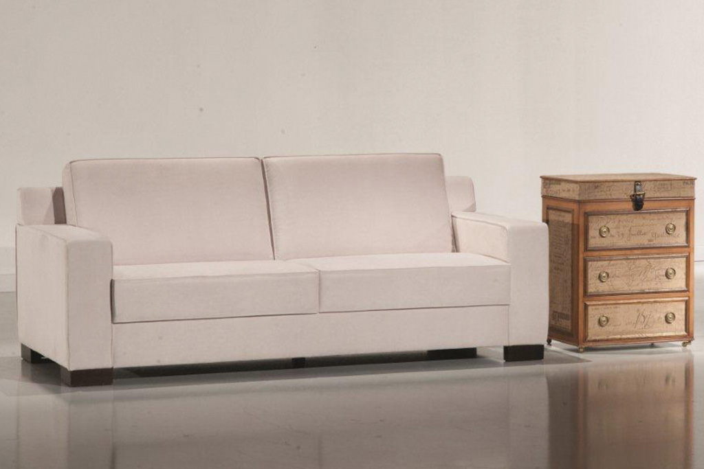 A imagem mostra um ambiente claro, com paredes brancas e um piso claro refletindo partes do sofá. No centro encontra-se o sofá na cor branca com quatro pés de madeira, um em cada canto do sofá. Na lateral direita do sofá encontra-se um móvel de madeira com três gavetas.