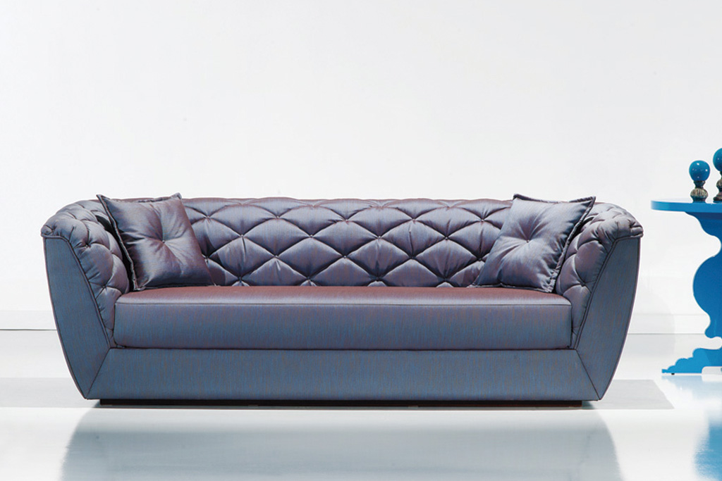 A imagem mostra um ambiente claro, com parede branca e piso branco. No centro encontra-se um sofá na cor azul com detalhes no estofado e dois travesseiros um em cada canto do sofá também nas cores azuis. Na lateral direita do sofá temos a metade de uma mesinha azul.